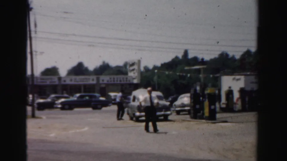 1957 Normal, Illinois street scene with men near a gas station, parked cars, storefront signs, and overhead wires.