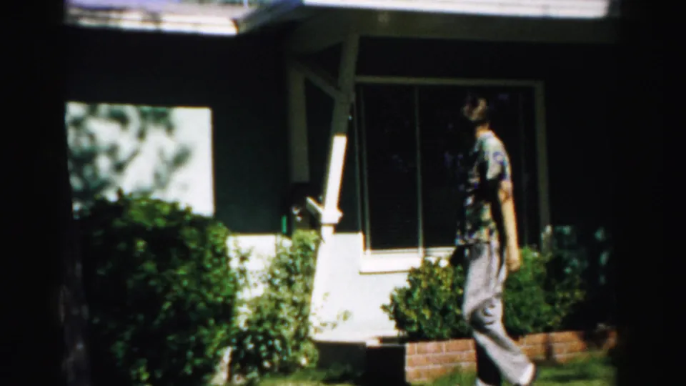 1957 Normal, Illinois home-movie frame of a man walking past a house exterior with shrubs, window, lawn, and strong shade.