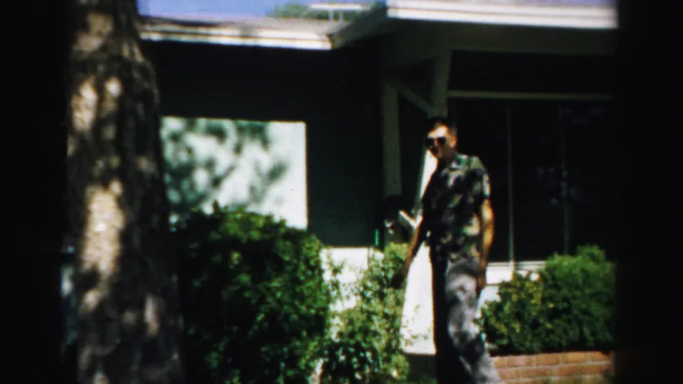 1957 Normal, Illinois frame showing a man in sunglasses near a house exterior with a window, shrubs, lawn, and tree trunk.