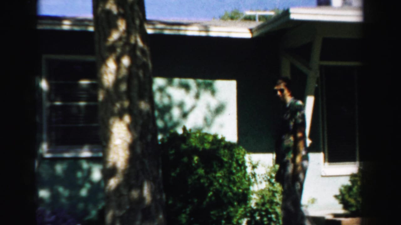 1957 Normal, Illinois 8mm frame showing a man outside a dark-sided house with shrubs, a window, and a tree trunk in front.
