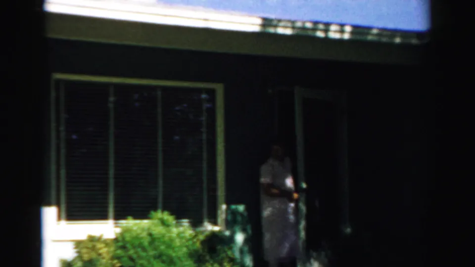 1957 Normal, Illinois 8mm frame showing a woman at a house doorway beside a large window and shrubs in bright sun and shadow.