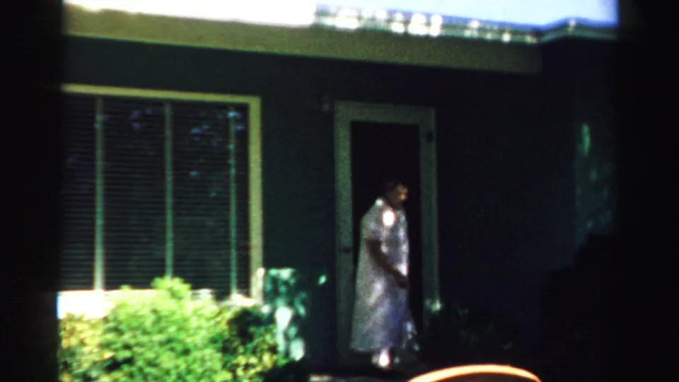 1957 Normal, Illinois 8mm frame showing a woman moving out from the front door near shrubs, window blinds, and the shaded house exterior.