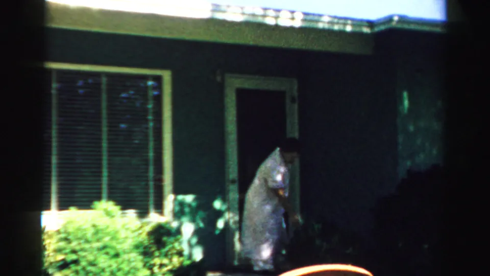 1957 Normal, Illinois 8mm frame showing a woman bending near the shrubs by the front doorway of a shaded residential exterior.