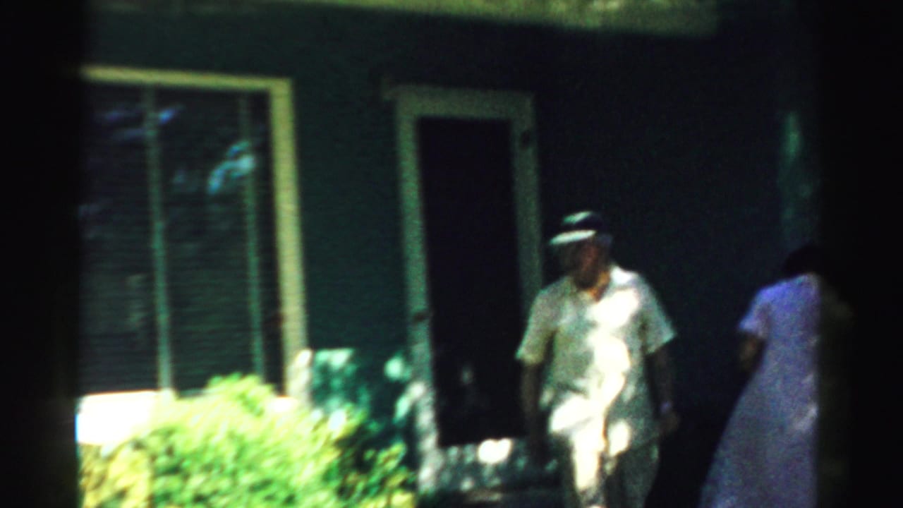 Older couple outside a dark house, with the man in a white visor near a doorway.