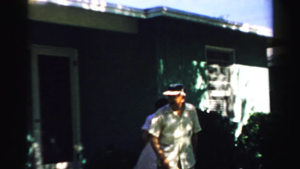 An older couple walks along the exterior of a house in Normal, Illinois, with strong sunlight and shade across the frame.