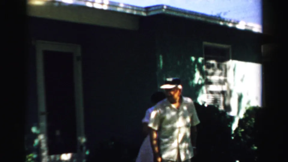 The older man walks near a house doorway while the woman is partly obscured behind him in grainy 8mm footage.