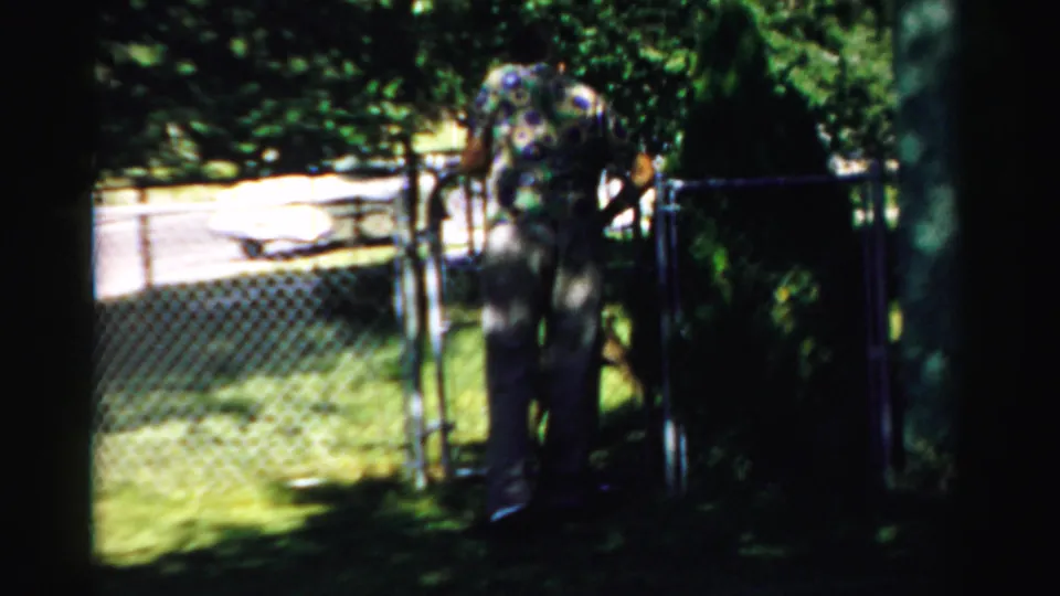 An older man in a patterned shirt and white visor appears near a chain-link gate in a sunlit residential yard.
