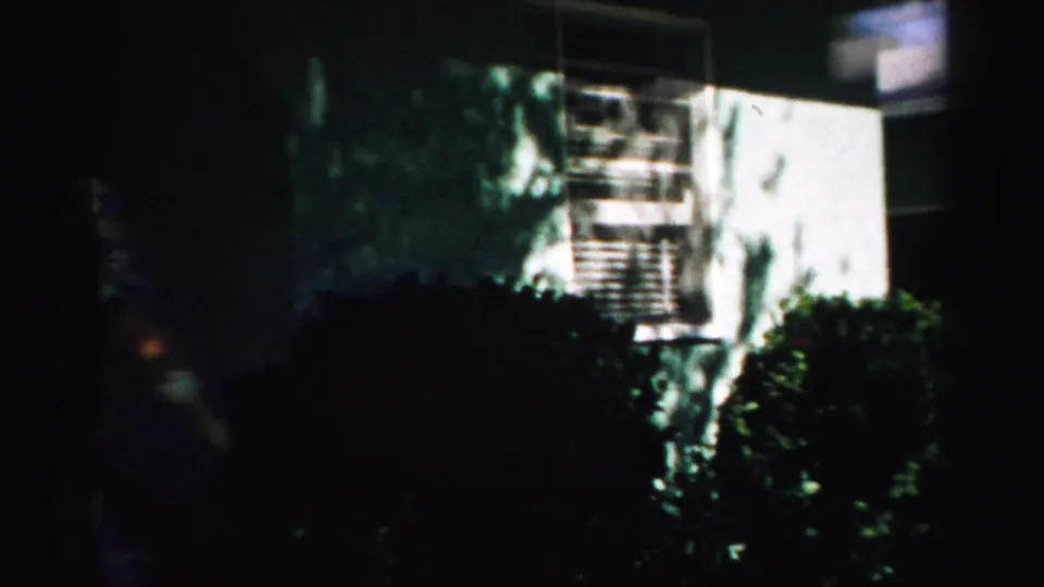 1957 Normal, Illinois home-movie view of a pale house wall, window, shrubs, and heavy foreground shadow.