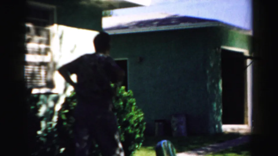 1957 Normal, Illinois frame showing a man silhouetted near shrubs beside a pale residential exterior and open doorway.