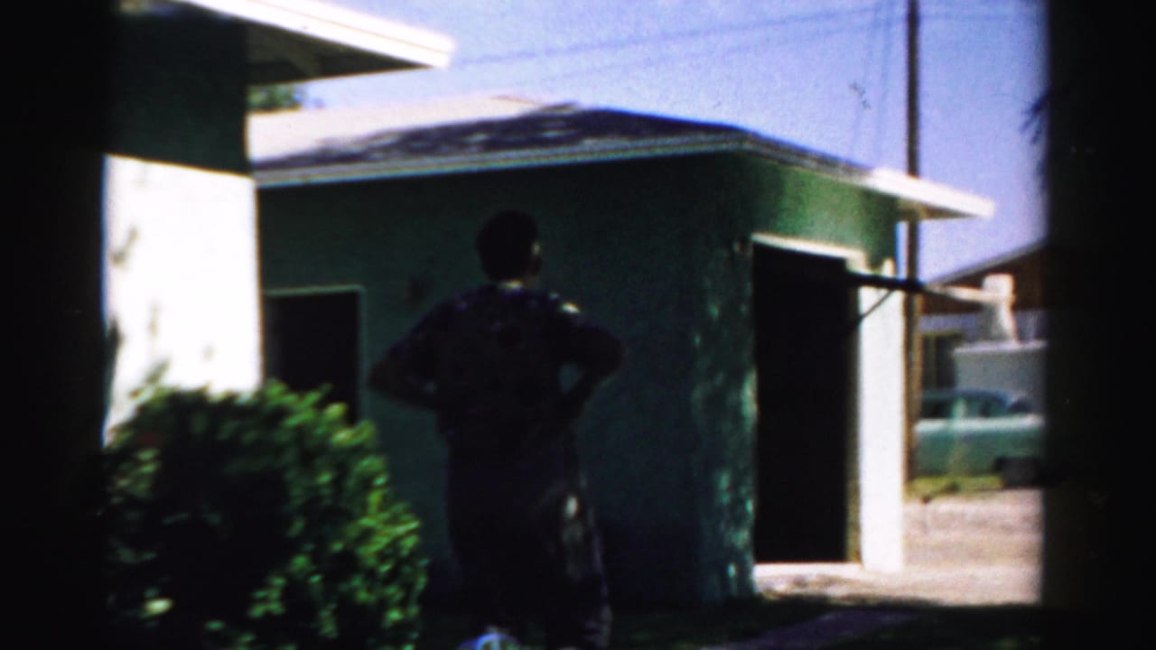 In Normal, Illinois, a man stands near an open garage at a green house, with lawn and driveway visible in 1957 home-movie footage.