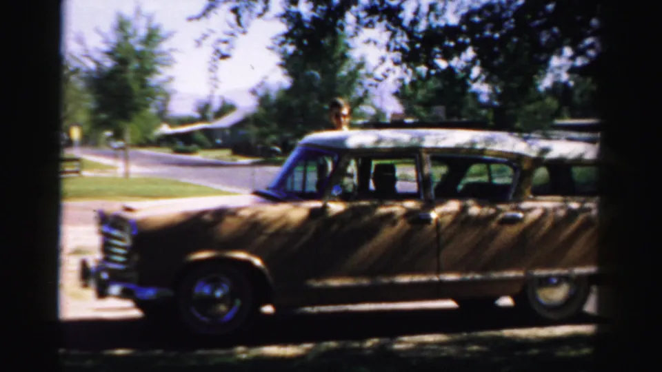 The station wagon is seen broadside near the street, with the man partly visible above or behind the car in the suburban setting.