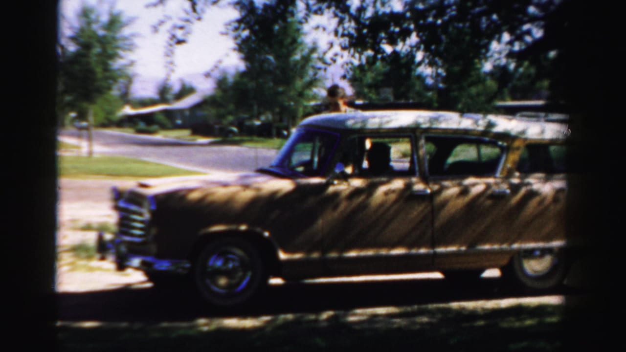 1957 Normal, Illinois home-movie frame showing a tan wood-paneled station wagon at the curb with an adult figure near the rear.