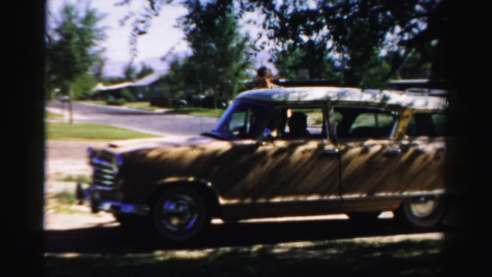 1957 Normal, Illinois home-movie frame showing a tan wood-paneled station wagon at the curb with an adult figure near the rear.