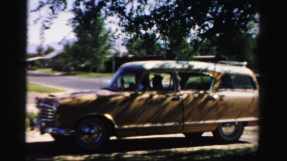 1957 Normal, Illinois frame with the tan station wagon framed by heavy shade above and a quiet residential street beyond.