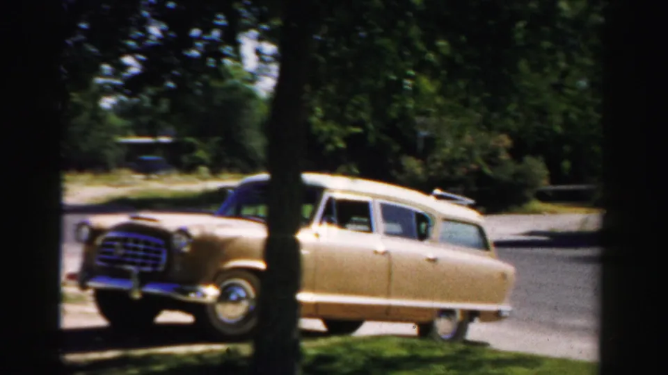 1957 Normal, Illinois frame of the station wagon angled on the street, with chrome front details visible between dark tree trunks.