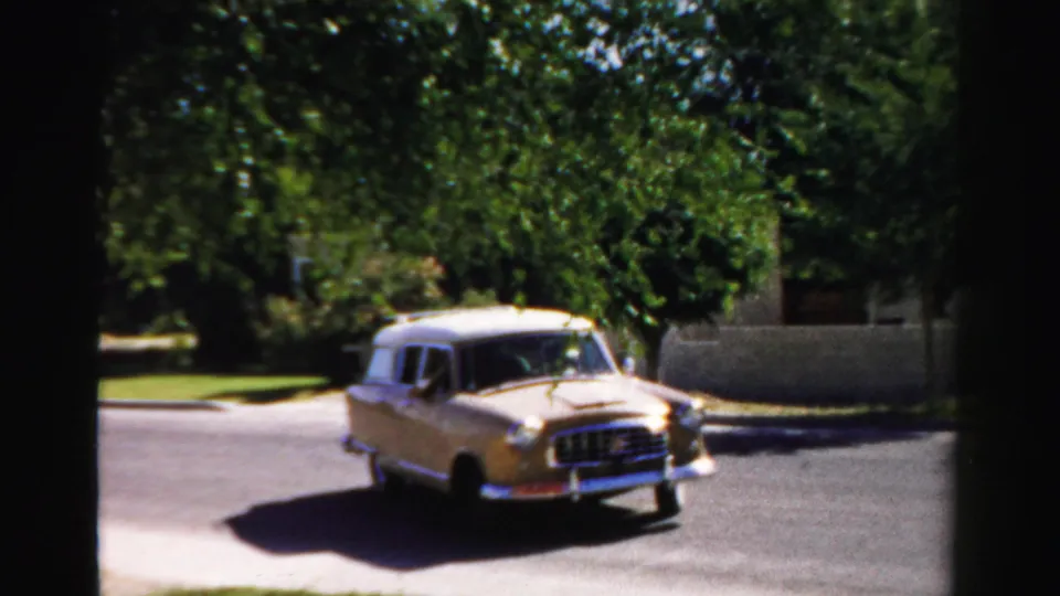 1957 Normal, Illinois frame showing the gold wagon angled in the street, casting a shadow near leafy yards and a low wall or fence.