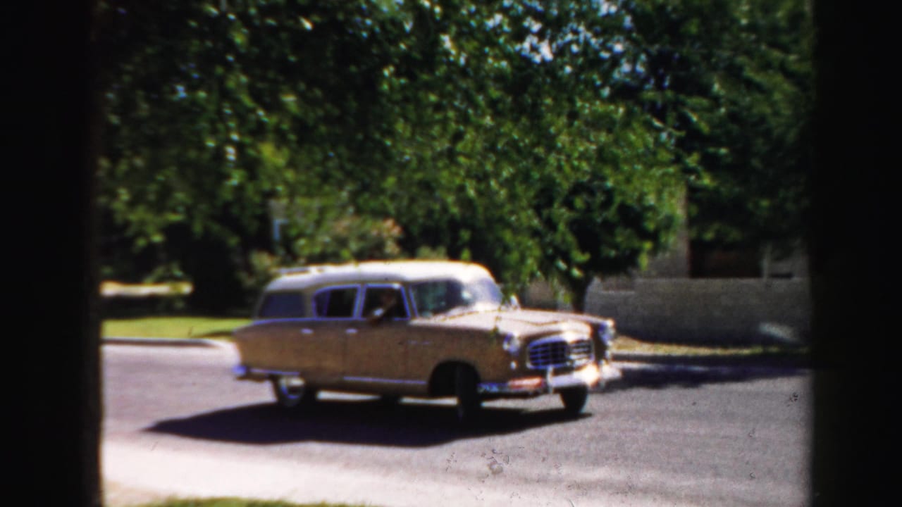 1957 8mm frame from Normal, Illinois showing a light-colored station wagon moving from a driveway or curb area onto a quiet street.