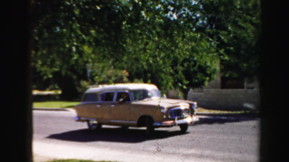 1957 8mm frame from Normal, Illinois showing a light-colored station wagon moving from a driveway or curb area onto a quiet street.