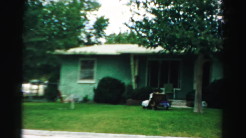 Home-movie view of a green single-story house in Normal, Illinois, with trees, shrubs, porch steps, and a bright lawn.
