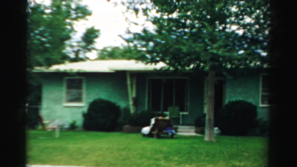 Final 1957 frame showing the green house front in Normal, Illinois, with lawn, shrubs, trees, and small items near the porch.