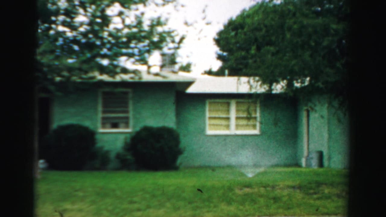 1957 Normal, Illinois 8mm frame of a teal single-story suburban house, front lawn, sprinkler spray, shrubs, and mature trees.