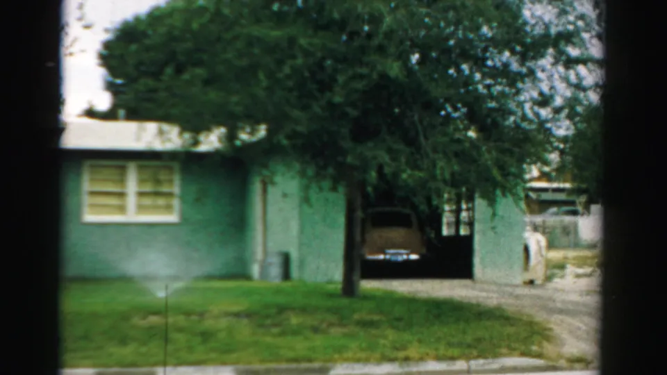 1957 Normal, Illinois frame showing the house, sprinklered lawn, large tree, gravel driveway, carport, and parked classic car.