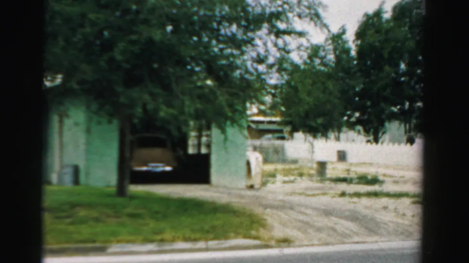1957 Normal, Illinois 8mm frame of a driveway and carport with a parked classic car, lawn edge, tree, and neighboring residence.