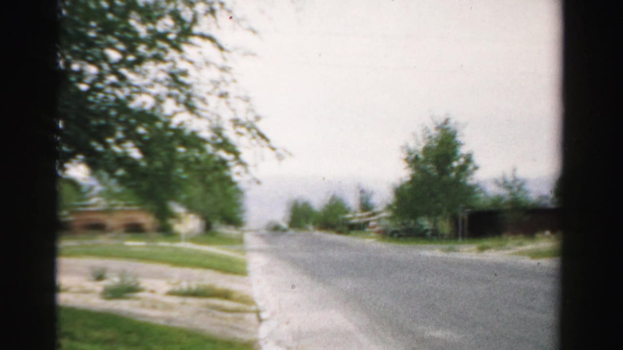1957 Normal, Illinois home-movie frame showing a quiet residential road with trees, lawns, and modest houses.