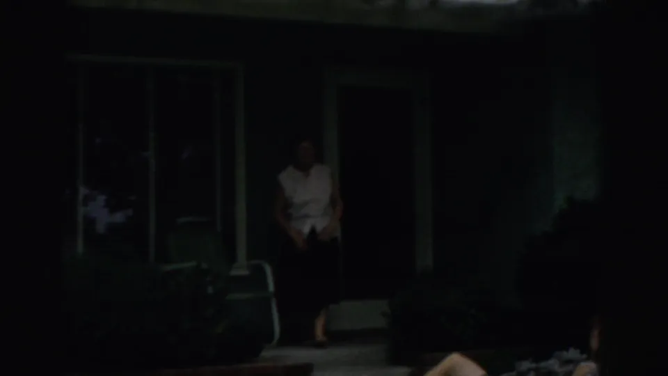 Dim 1957 porch frame showing a woman near a front doorway, with a window, chair, and shaded house exterior.