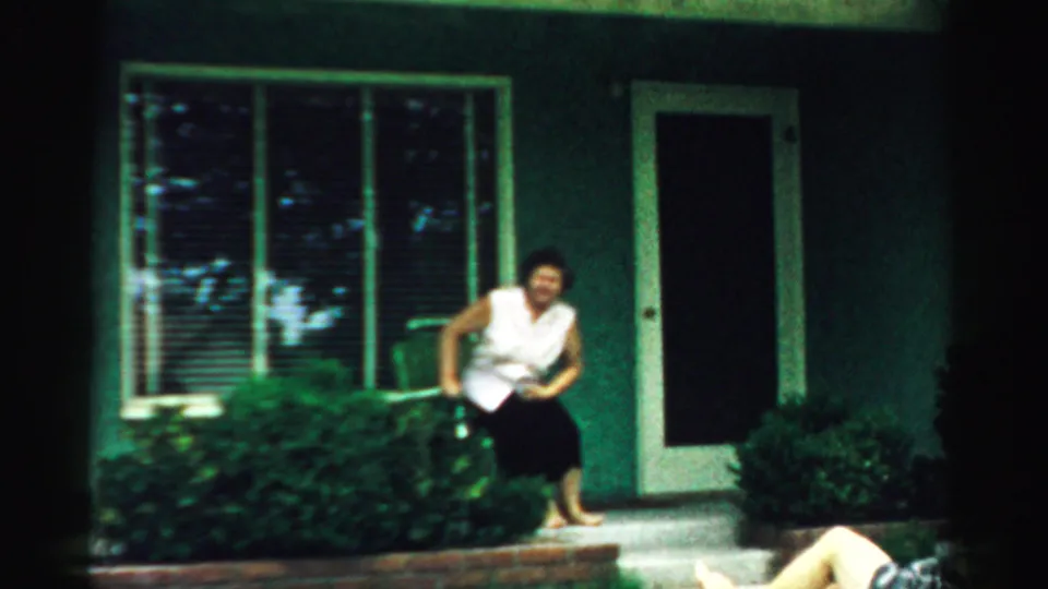 1957 Normal, Illinois frame of a woman in a white top and dark skirt seated on porch steps by a window and shrubs.