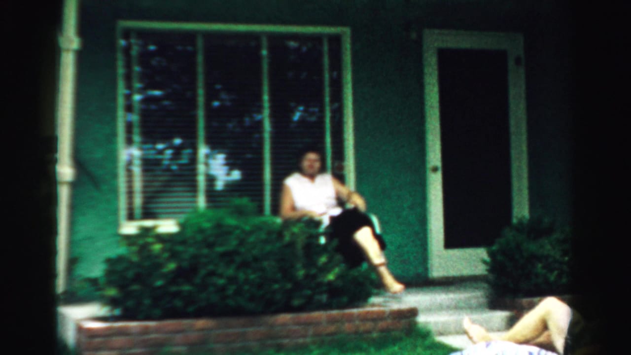 Woman seated on front steps beside shrubs at a green house.