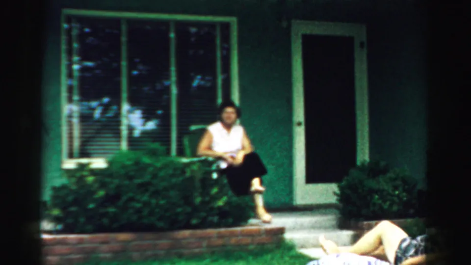 A 1957 Normal, Illinois frame shows a woman seated near a house doorway with shrubs below the window and a person partly visible on the grass.