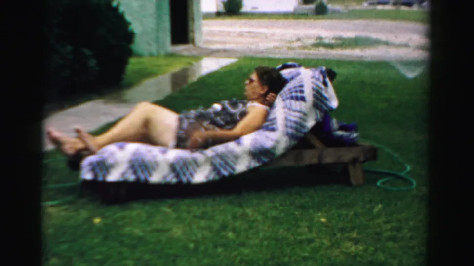 In 1957 Normal, Illinois home-movie footage, a woman reclines on patterned lawn furniture beside a sidewalk and garden hose.