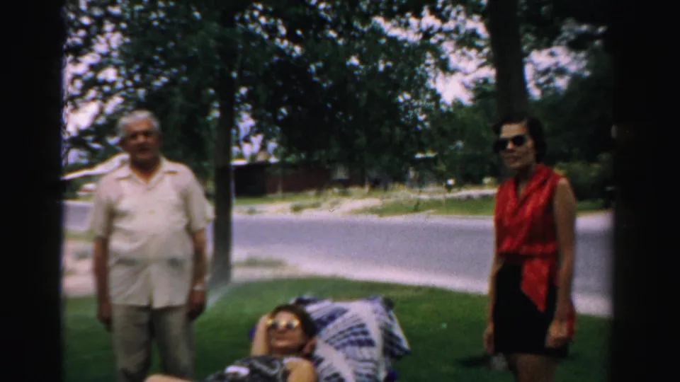 An elderly man and a woman in sunglasses stand beside a reclining woman on a lawn chair in 1957 Normal, Illinois footage.