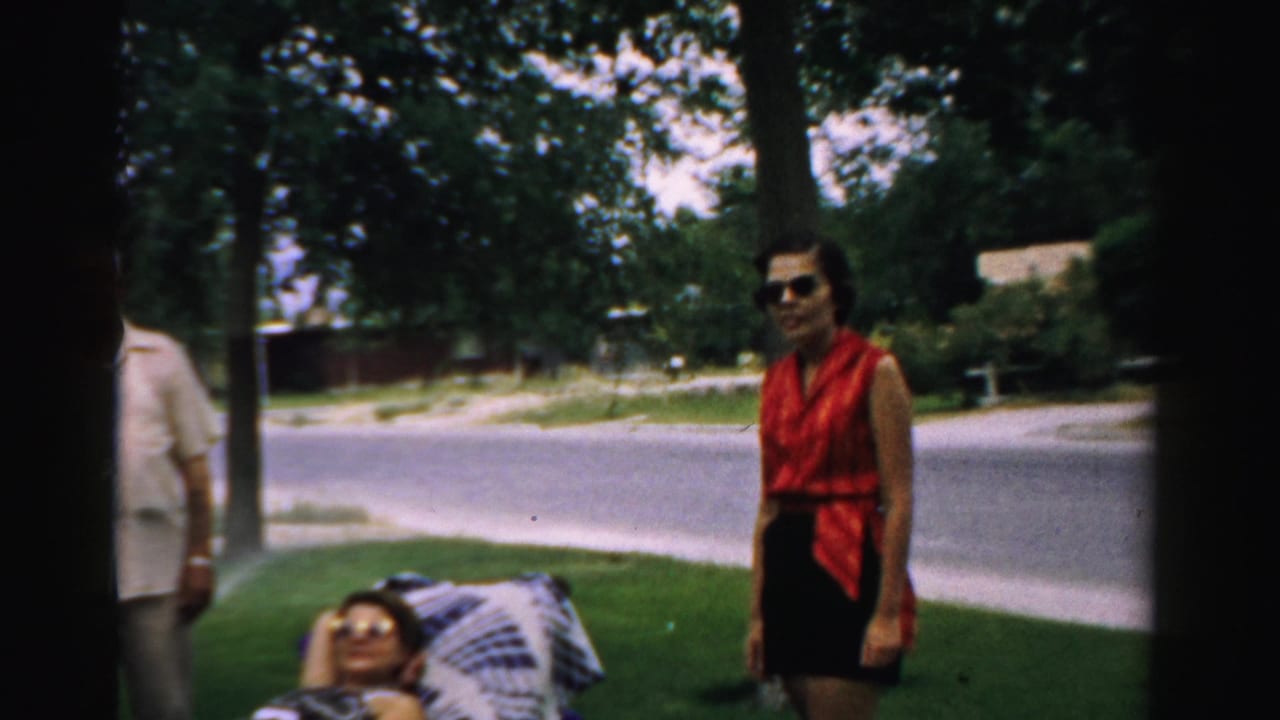 1957 Normal, Illinois home-movie frame showing adults in a yard, a woman in a red sleeveless top, and another person reclining near a quiet street.