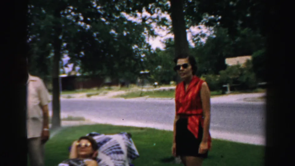 1957 Normal, Illinois home-movie frame showing adults in a yard, a woman in a red sleeveless top, and another person reclining near a quiet street.