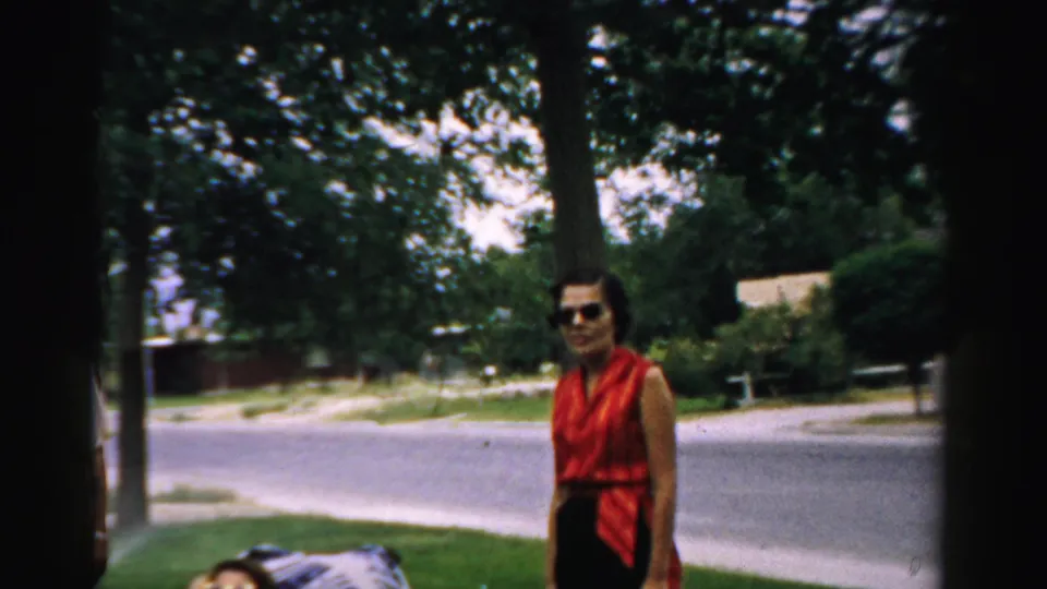 1957 Normal, Illinois yard scene with a woman in sunglasses standing under trees, with grass, street, and houses in the background.