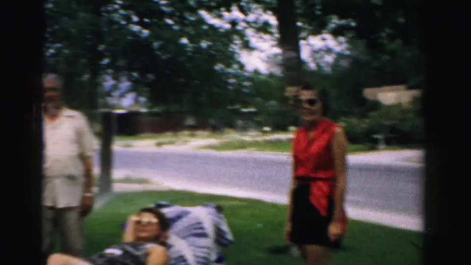 Home-movie frame from 1957 Normal, Illinois showing adults gathered in a shaded yard, including a standing man, a standing woman, and a reclining adult.