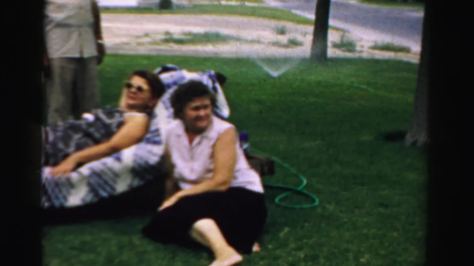 1957 Normal, Illinois yard frame with two adults relaxing on grass near a patterned blanket, a green garden hose, trees, and a residential street.