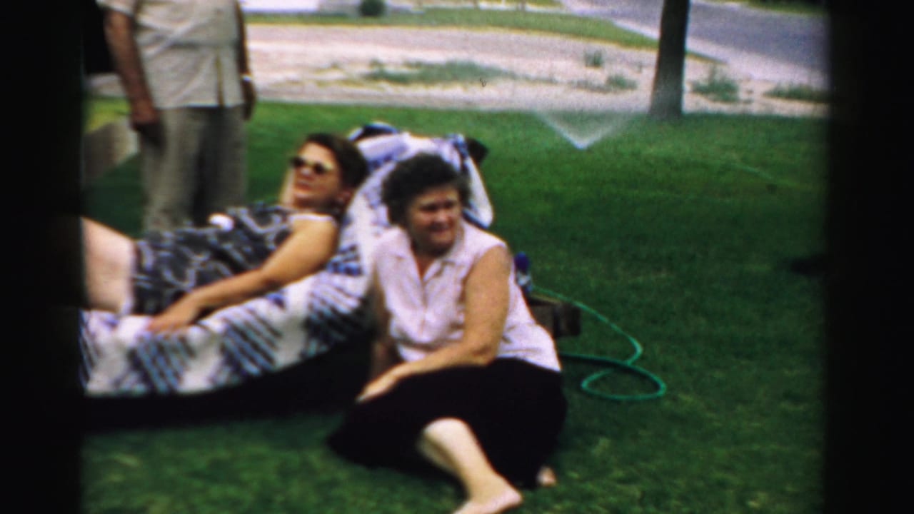 Two women relax on a patterned lawn chair while a man stands nearby on a residential lawn.
