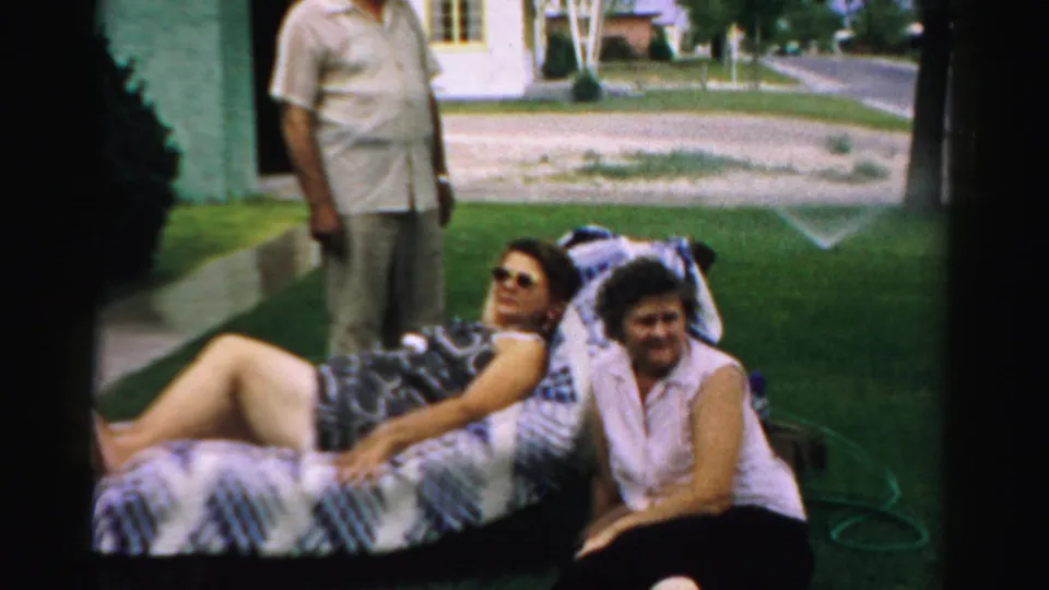 Two women relax on a front-lawn chair in Normal, Illinois, with a man standing nearby and residential houses in the background.
