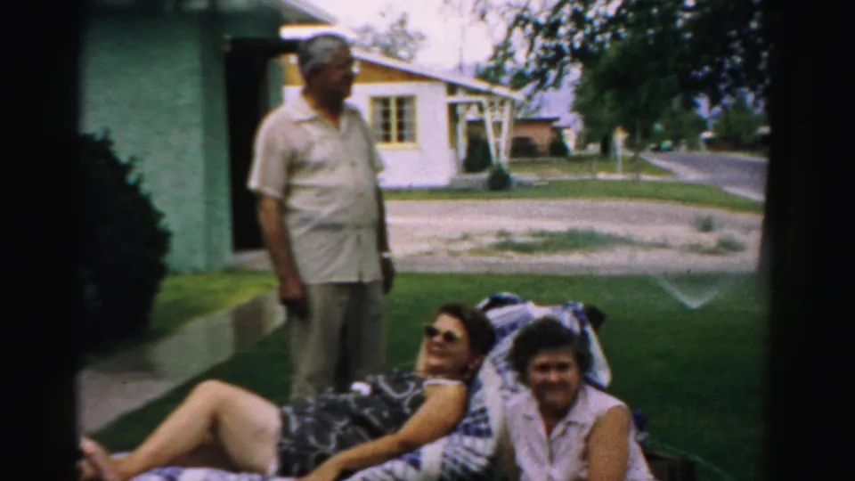 A standing man and two women on a lawn chair appear in a 1957 Normal, Illinois home-movie view of a suburban front yard.