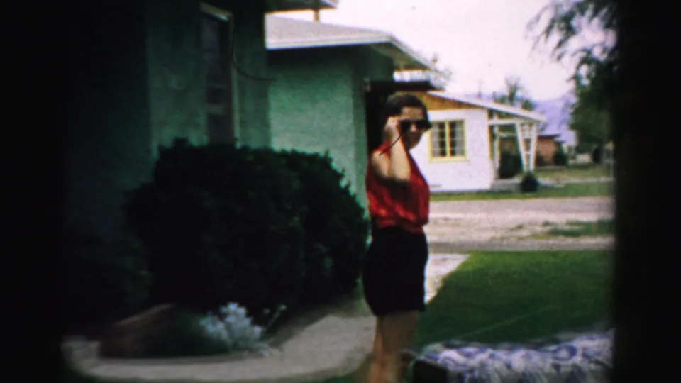 A woman in a red top appears to hold a camera on a suburban lawn in Normal, Illinois, with houses and landscaping behind her.