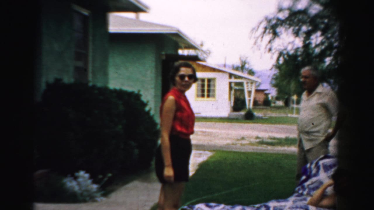 Woman in red sleeveless top stands by a house while an older man stands on the lawn.
