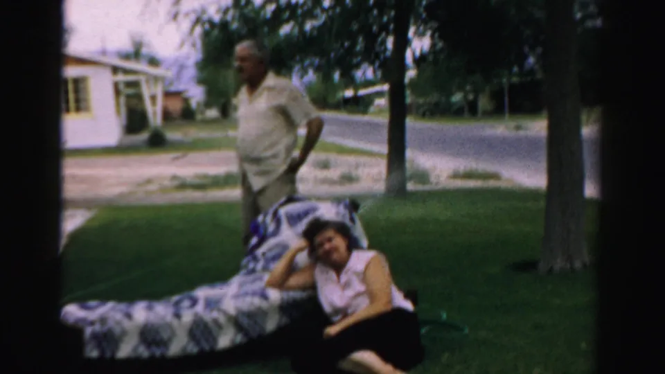 In the 1957 Normal, Illinois yard scene, an older man stands near a woman seated on grass beside a patterned blanket or lounge.