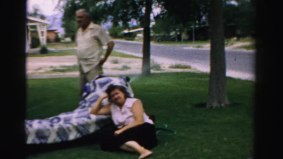The 1957 Normal, Illinois home movie shows an older man standing while a woman sits barefoot beside a patterned blanket in a suburban yard.