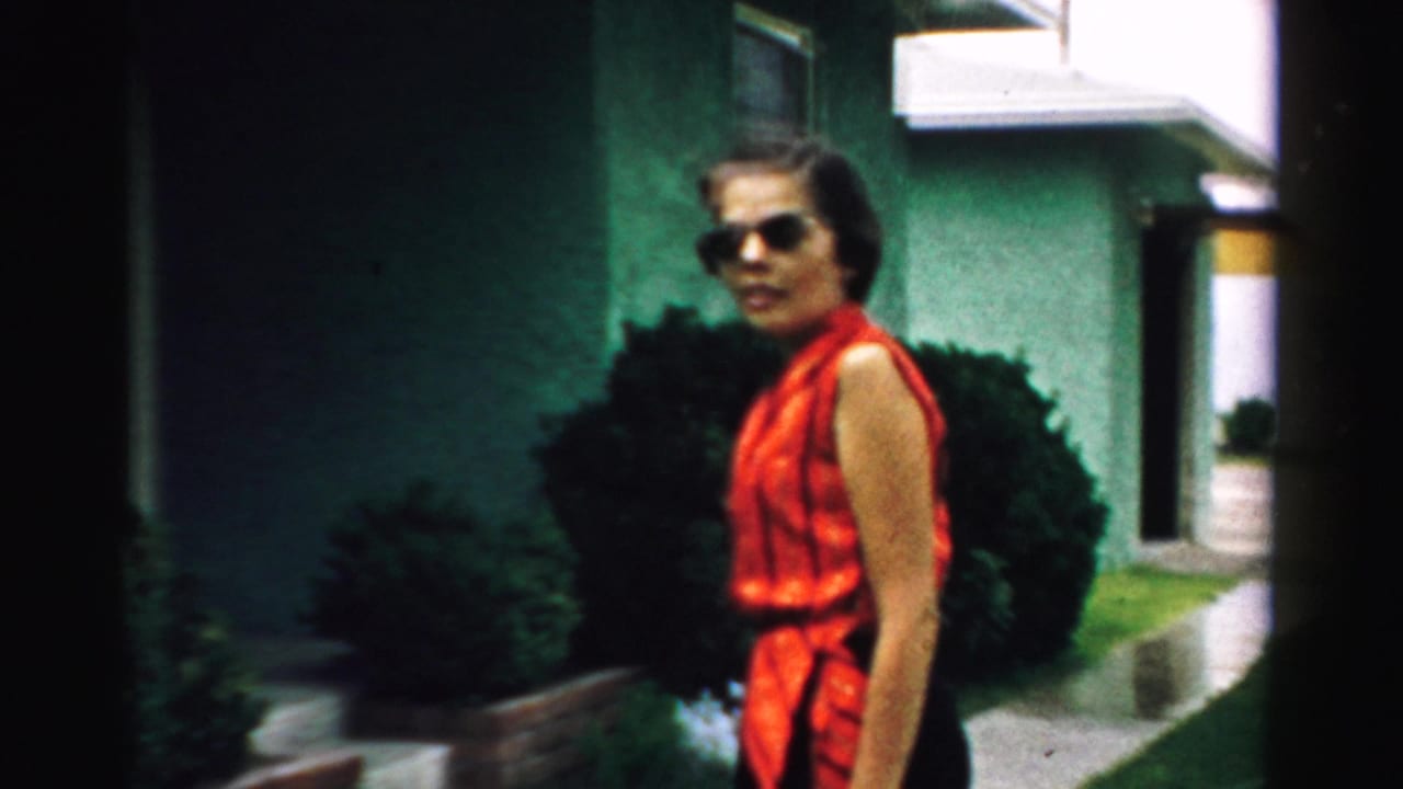 In Normal, Illinois home-movie footage from 1957, a woman in a red sleeveless top walks past shrubs and a green stucco house.