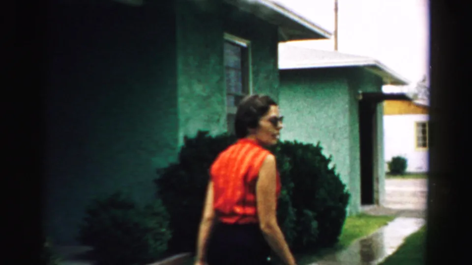 A woman in a red sleeveless top walks away across a suburban yard in Normal, Illinois, with green houses and shrubs behind her.