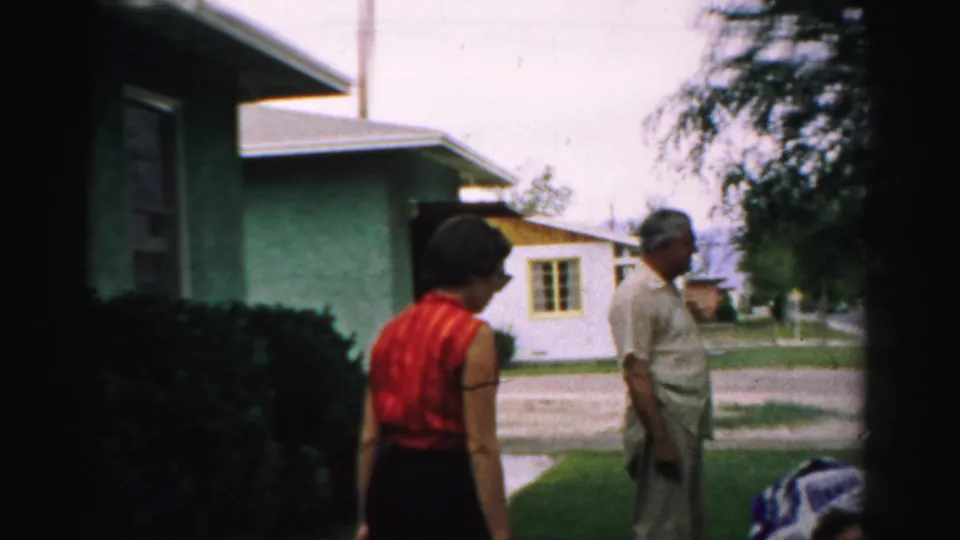 The woman in red approaches a standing man in a 1957 Normal, Illinois yard, with neighboring homes and trees visible.