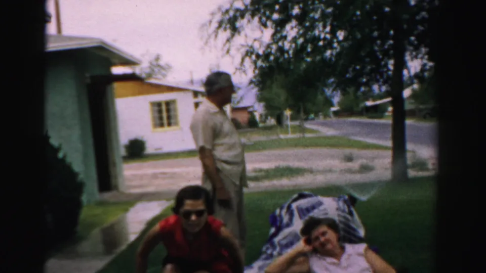 A man stands in a suburban yard while one woman in red sits and another reclines on a blanket, with houses and fencing nearby.